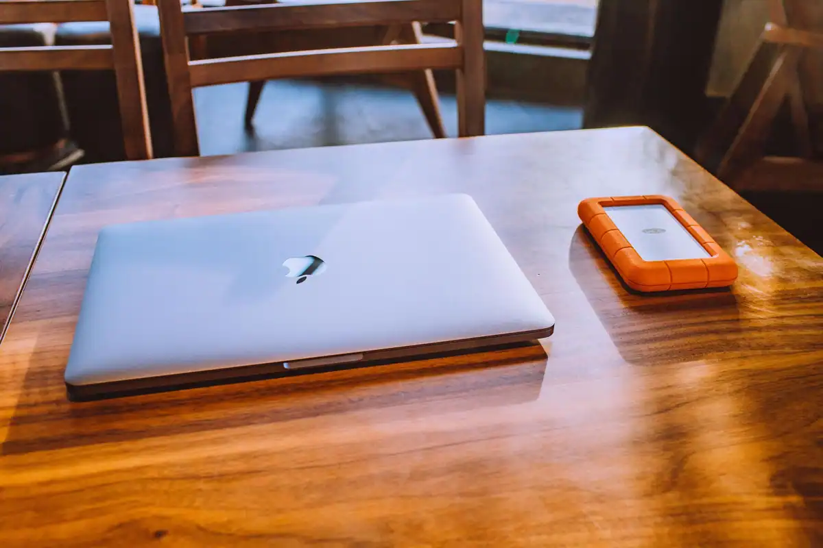 Laptop închis pe o masă din lemn, lângă un hard disk extern portocaliu
