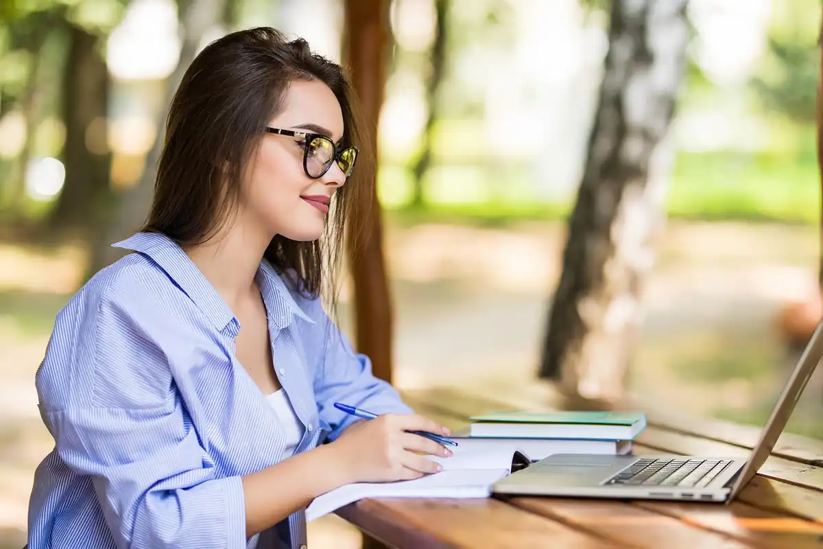 Studentă care scrie într-un caiet, cu laptopul deschis pe o masă în aer liber
