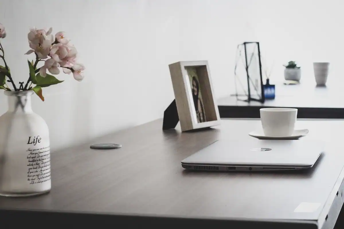 Birou ordonat cu laptop închis, ramă foto, vază cu flori și o ceașcă