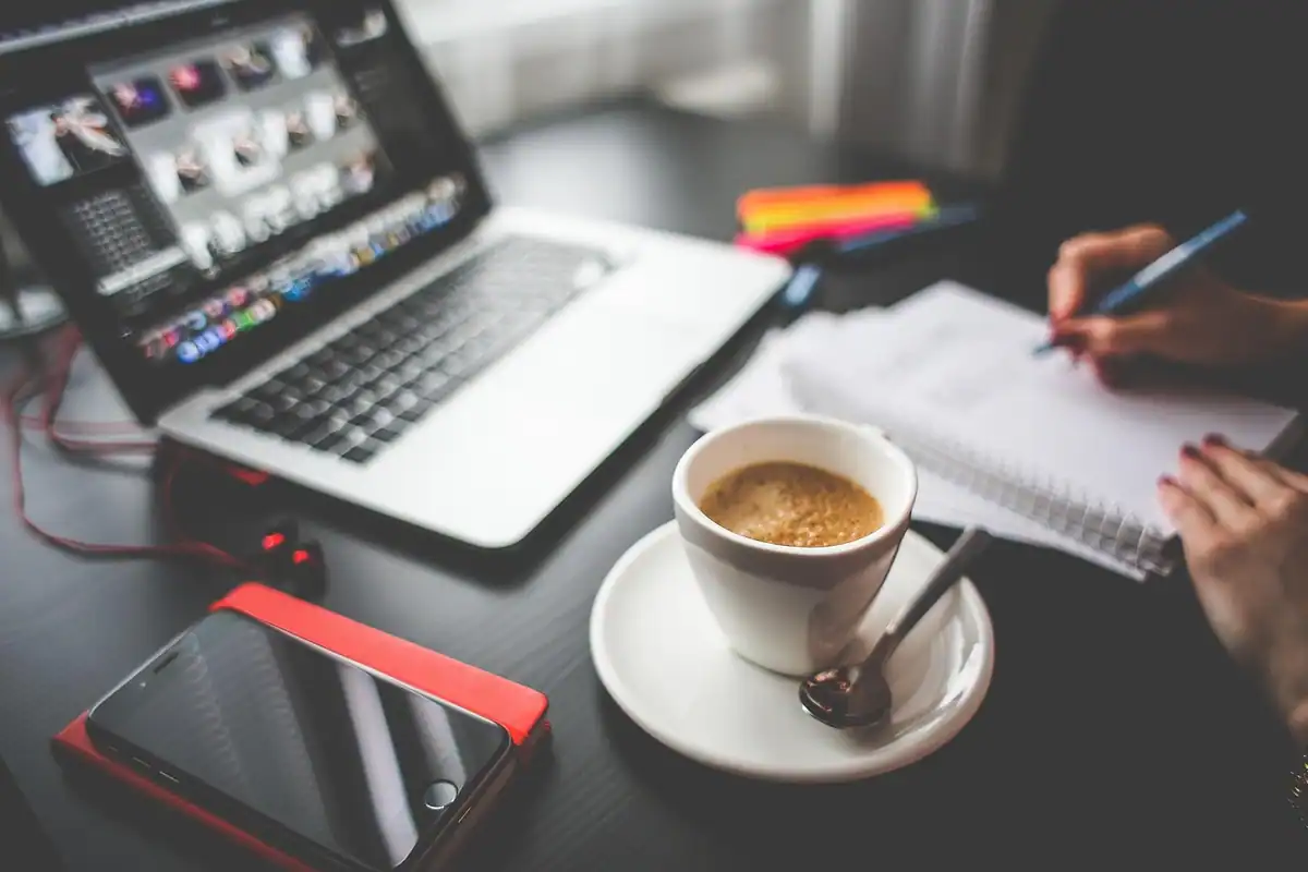 Persoană care notează într-un caiet, lângă un laptop, o cafea și un telefon
