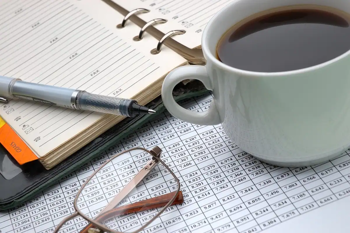 Agendă cu inele, cană de cafea, pix și ochelari așezate pe o foaie cu tabel