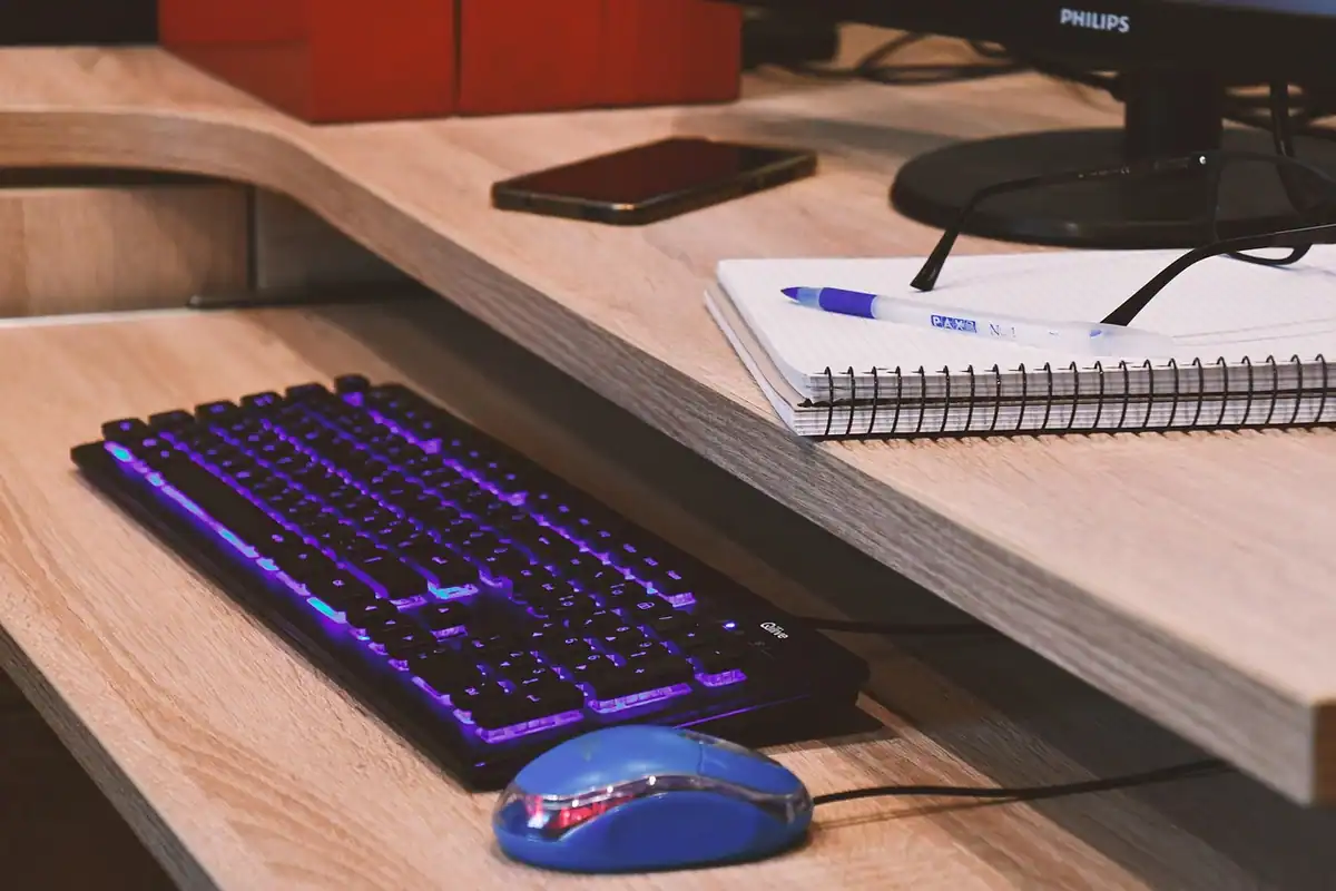 Tastatură luminată violet, mouse albastru și caiet cu spirală pe un birou din lemn