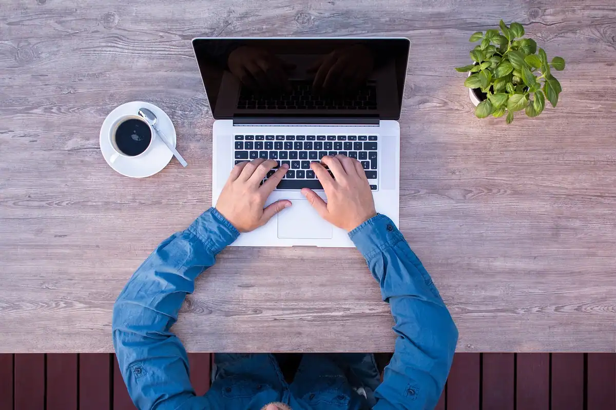 Persoană în cămașă albastră care scrie pe un laptop, văzută de sus, cu o cafea alături