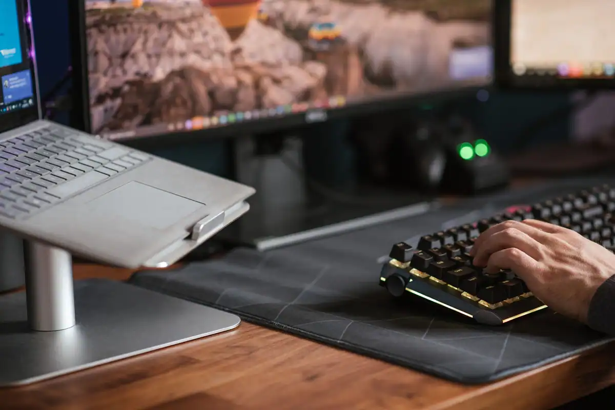 Mâini pe o tastatură aprinsă, în fața unui laptop pus pe suport și a unui monitor
