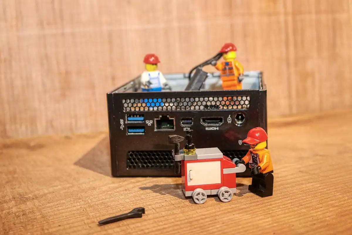 Mini PC negru compact, cu porturi vizibile, înconjurat de mici figurine de muncitori