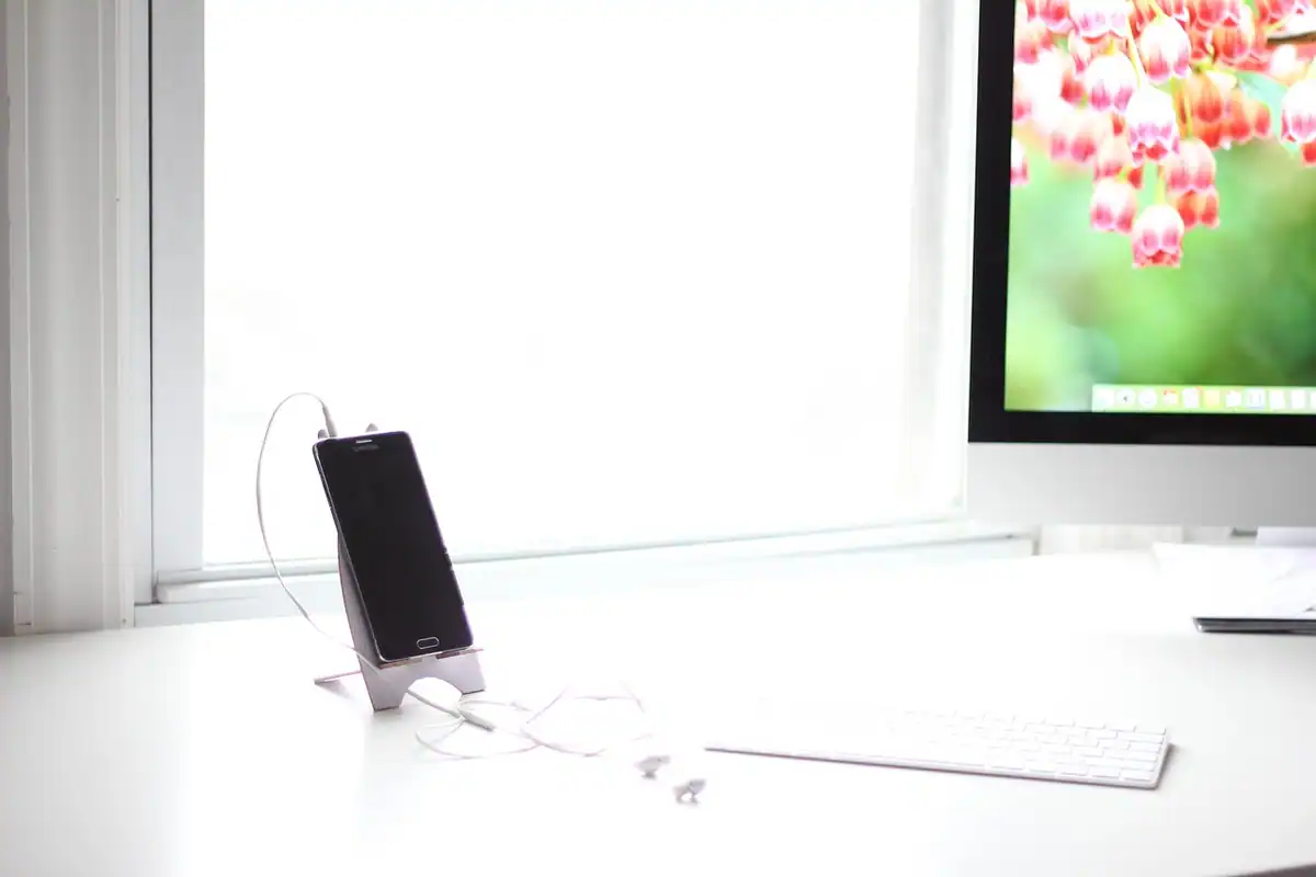 Telefon așezat pe un suport pe un birou alb, lângă un monitor cu imagine florală