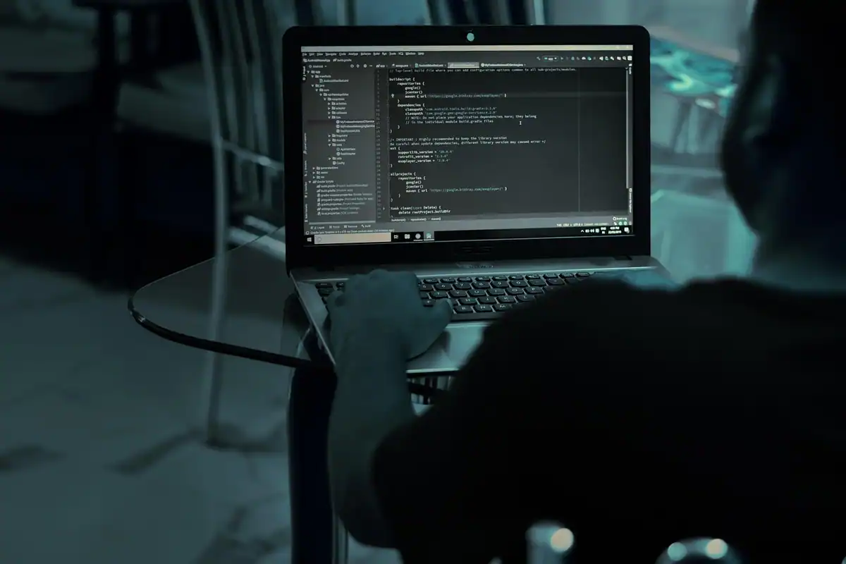 Silueta unei persoane cu laptopul deschis, pe care se vede cod, într-o cameră întunecată
