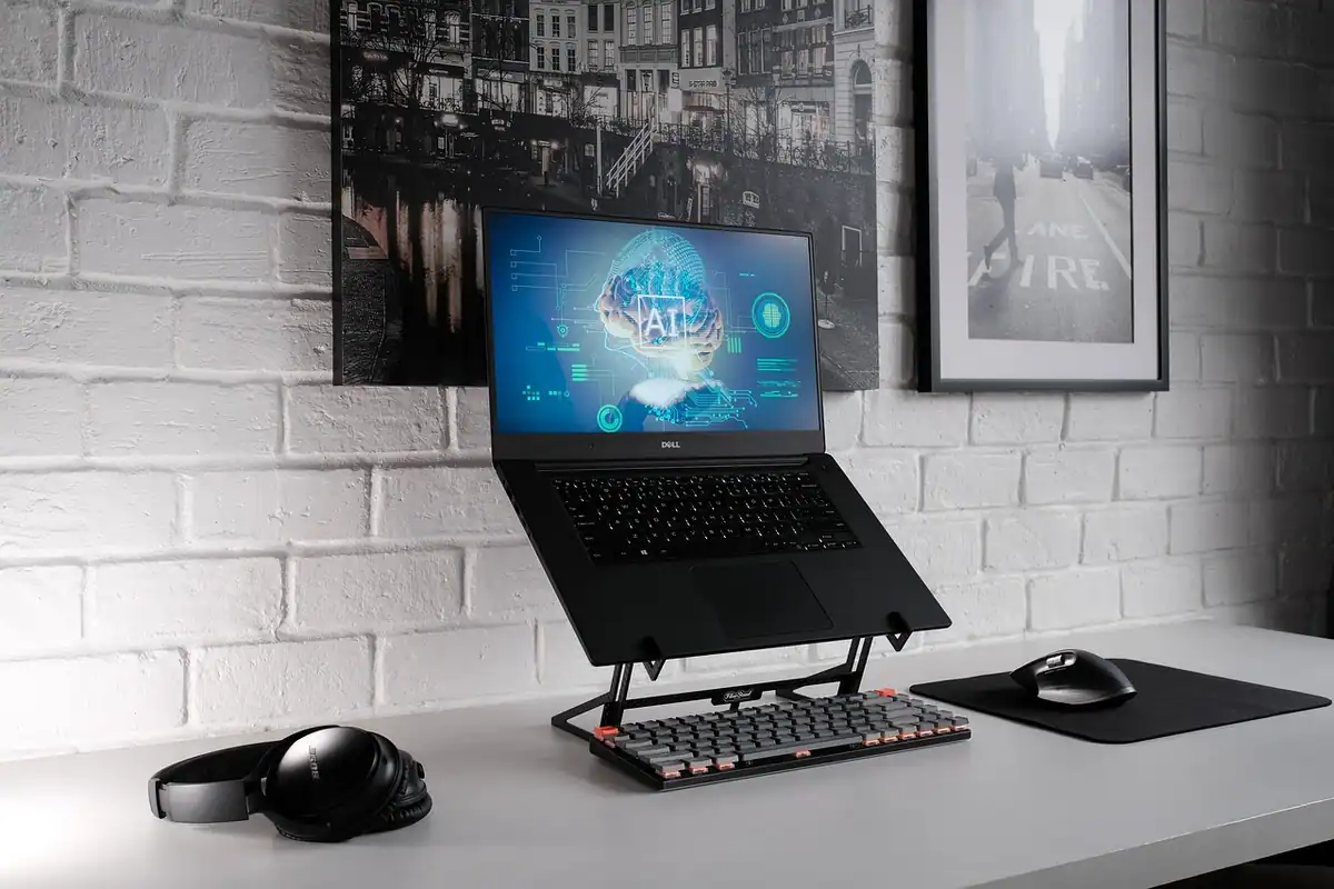 Laptop pe suport metalic, lângă mouse și căști, pe un birou alb