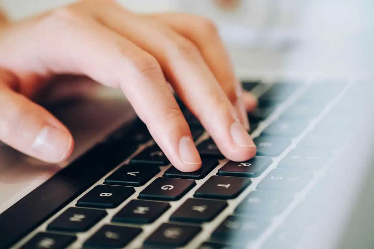 Mâini care scriu pe tastatura unui laptop, în lumină blândă de zi