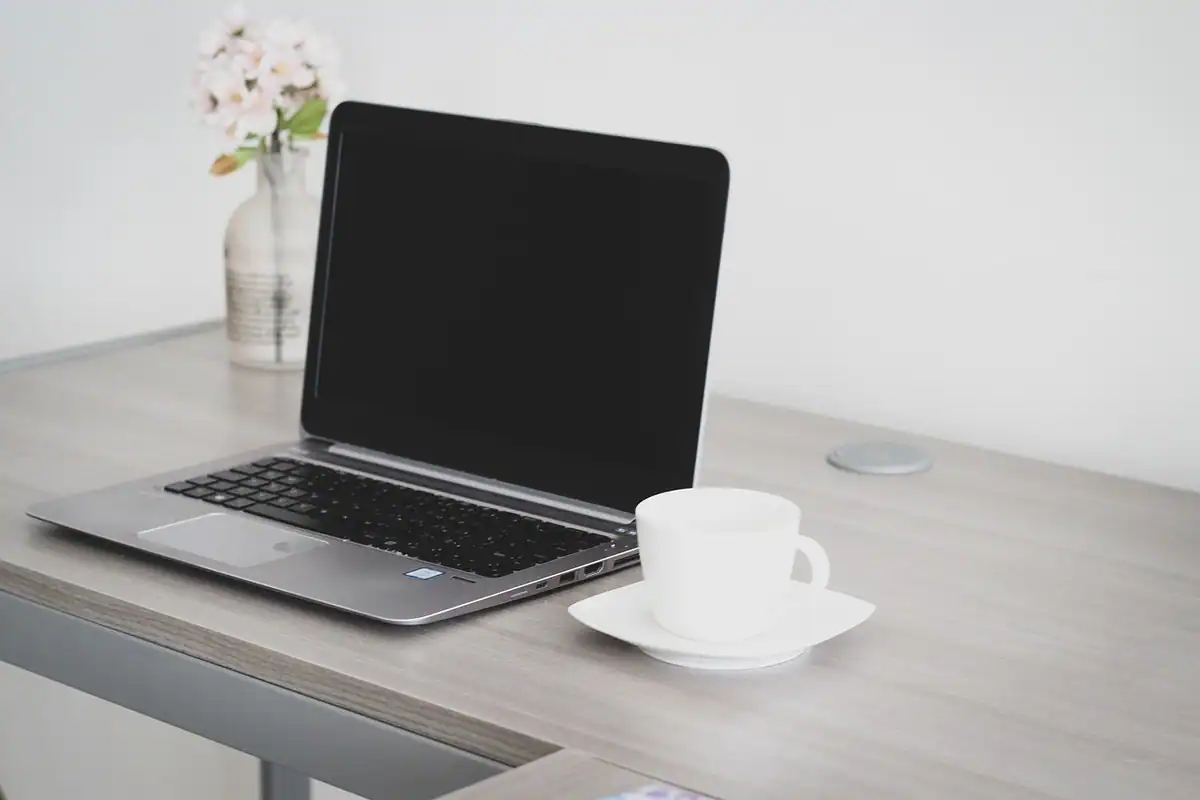 Laptop deschis pe un birou de acasă, lângă o ceașcă albă și o vază cu flori